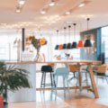 Ein Coworking-Space mit mehreren Schreibtischen, Pflanzen und großen Fenstern, die Tageslicht in den Raum lassen. Menschen arbeiten an Laptops.